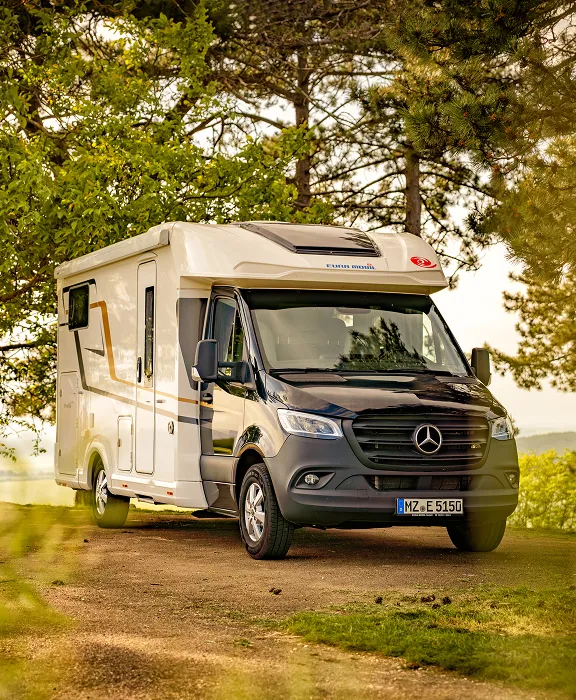 Teilintegriertes Mercedes-Wohnmobil auf einem sonnigen Stellplatz unter grünen Bäumen im Wald.